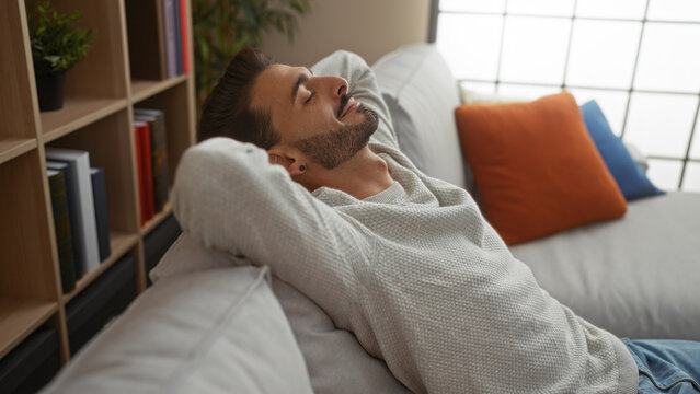 Young man relaxing indoors on a couch in a cozy living room, showcasing a handsome, attractive appearance with a contented expression in a modern apartment setting.