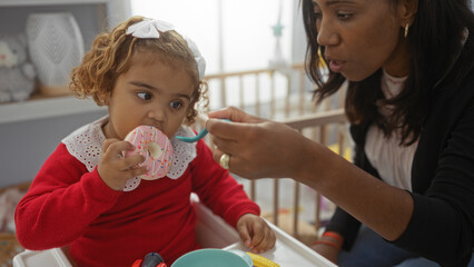 Woman feeding young daughter sitting with colorful toy donut in cozy home nursery fostering family connection indoors.