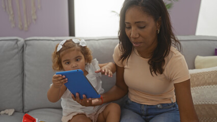 Woman and daughter using tablet indoors in living room, showcasing family bond, warm interaction, and cozy home environment with wooden decor and pastel colors.