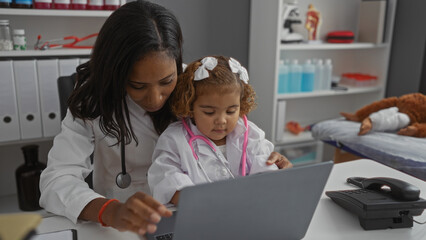 Obraz premium Woman doctor with a young girl wearing a pink stethoscope in a clinic room using a laptop surrounded by medical equipment and paperwork in a supportive healthcare environment.