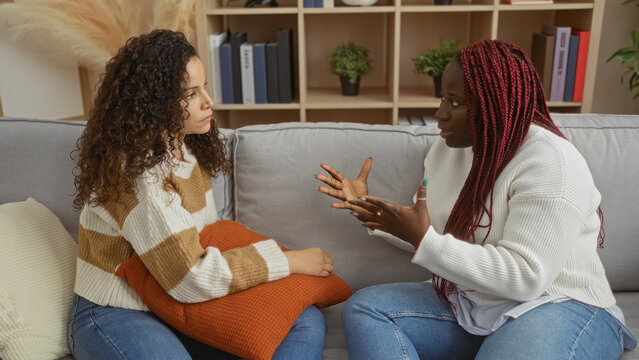 Two women friends sitting in a cozy living room engaged in a serious conversation, emphasizing their strong bond and emotional connection within a comfortable home setting.