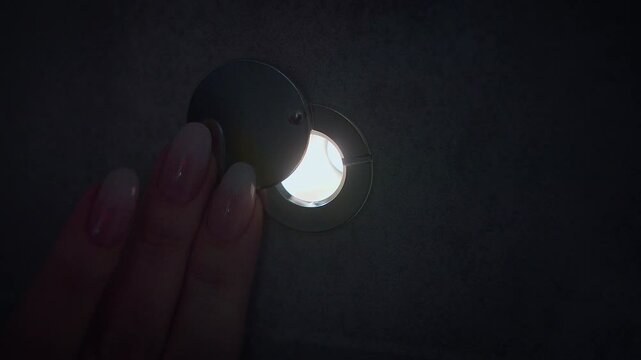 In the dark, women's fingers move the shutter of the door peephole, behind the door there is a bright light, a close-up