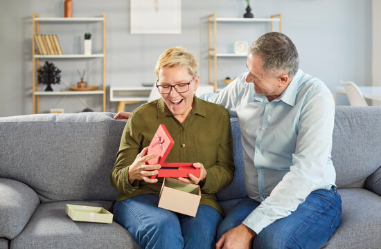 Portrait of happy smiling senior couple sitting on sofa in living room at home with present gift box celebrating marriage anniversary or birthday holiday. Love and relationships concept. - Powered by Adobe