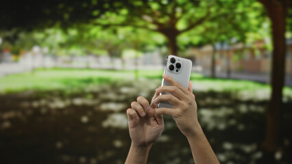 Hands holding smartphone in sunny outdoor park setting, capturing nature's beauty amidst lush green...