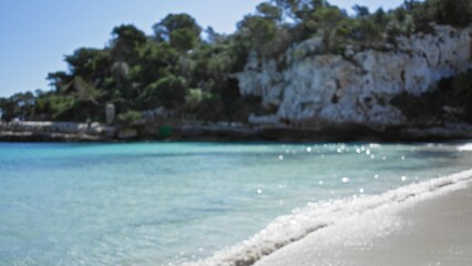 Blurred beach scene at cala llombards, mallorca with defocused turquoise water and bokeh effect on sandy shore on a sunny day.