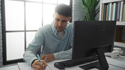 Man working indoors in office with computer and plant, focused on writing at a modern desk,...