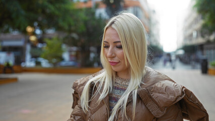 Obraz premium Woman standing outdoors in an urban park setting, showcasing beautiful blonde hair and wearing a brown jacket on a city street with a soft, calm expression.