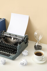 Vintage typewriter with crumpled paper, cup of coffee and hourglass on table near beige wall