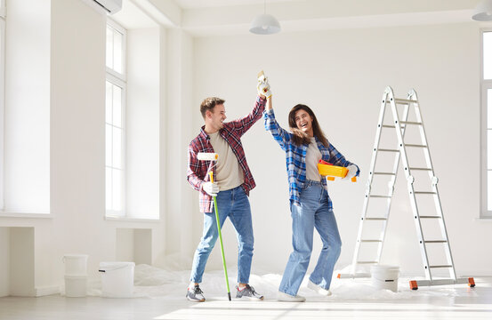Funny happy young couple having fun and dancing while painting the wall of their new apartment holding paint rollers. Married man and woman doing renovation. Moving and repair concept.