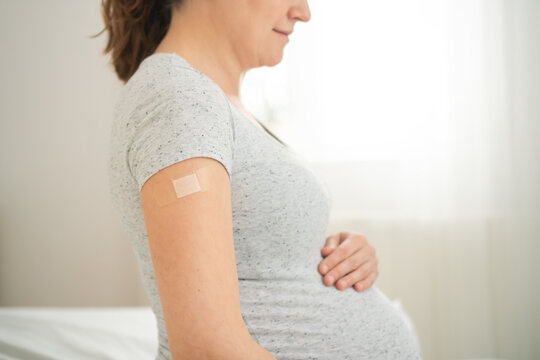 Image of a pregnant woman holding their upper arm with a bandage applied after receiving a vaccination. The scene emphasizes the importance of health care and protection.