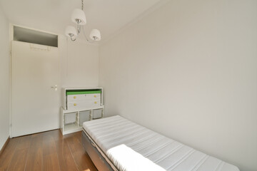 A bright and minimalist bedroom featuring a single mattress on a wooden floor, complemented by a modern chandelier and a compact storage unit.