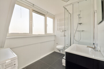 A sleek modern bathroom design featuring a glass shower, toilet, and washing machine, accentuated by ample natural light from large windows.