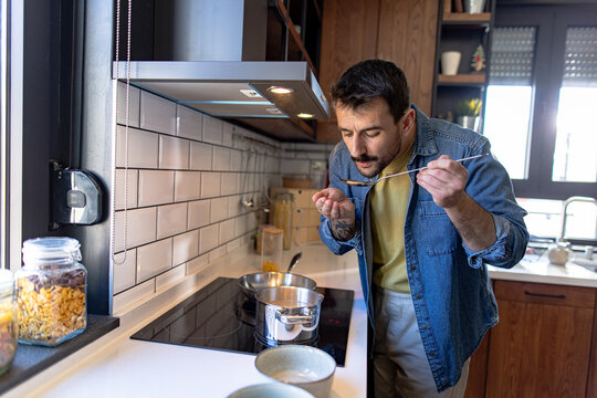 Man tasting food from a spoon while preparing a meal in a sleek, modern kitchen, surrounded by the aromas of delicious flavors