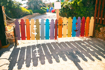 Small colorful wooden gate made of vertical planks painted in different bright colors, some featuring anchor cutouts. Sunlight casts long, distinct shadows of the gate on the cobblestone path below