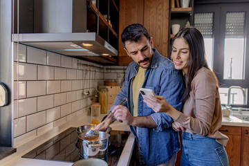 Young couple cooking together in a modern kitchen, using a smartphone to follow a recipe and create...