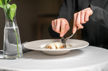 A close-up of a hand using a fork and knife to cut into a gourmet dish, elegantly presented on a white plate. A glass vase with greenery adds charm to the upscale dining atmosphere.
