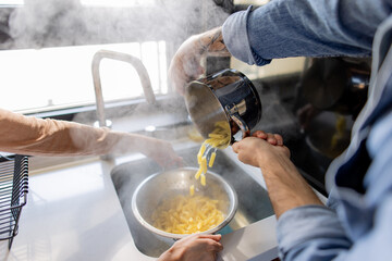 Two chefs skillfully draining steaming pasta in a colander, showcasing teamwork in a bustling professional kitchen environment