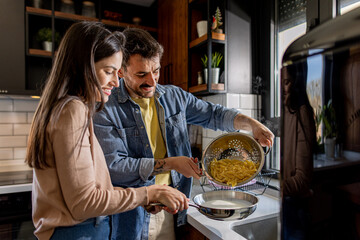 Happy couple enjoying quality time in their modern kitchen, preparing pasta and skillfully draining...