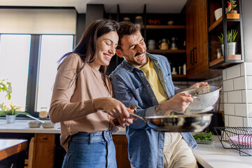 Young couple cooking together in a modern kitchen, laughing and enjoying the process of preparing delicious pasta and fresh vegetables