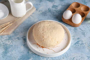 Plate with fresh raw dough, wheat and wooden holder of eggs on blue background