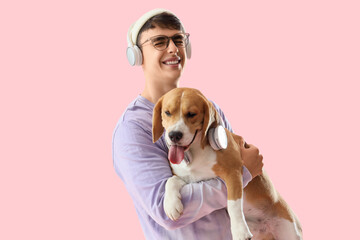 Young man in eyeglasses and headphones holding adorable Beagle dog on pink background
