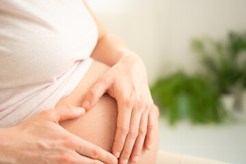 Close-up of a pregnant woman gently holding her belly, forming a heart shape with her hands. The image conveys a sense of love, anticipation, and maternal connection.