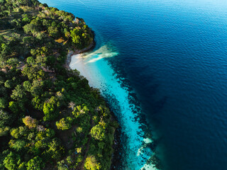 Tropical forest meets a white sandy beach with turquoise water along a curved island coastline, seen from above in Thailand.