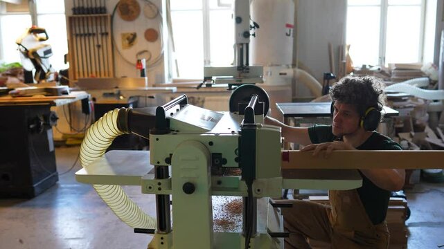 Joiner artisan in protective earmuffs adjusts twists jointer machine to shape wooden beam to desired thickness in workshop. Carpenter making furniture, craftsmanship and manual production business