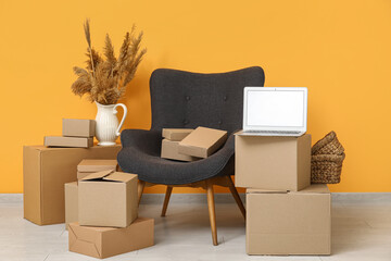 Blank laptop with moving boxes, armchair and pampas grass near orange wall in room