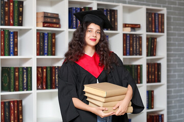 Female graduating student with stack of books near bookshelves in library