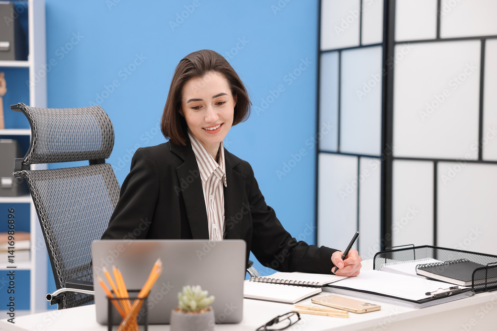 Poster smiling secretary taking notes while working on laptop at table in office
