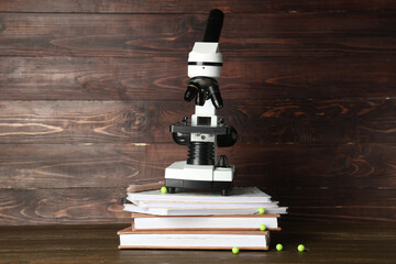 Modern microscope with books and pins on table against wooden background