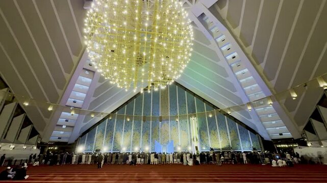 Interior of Shah Faisal Masjid, Islamabad with shiny yellow chandelier, red carpet, and prayers
