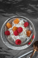Healthy Breakfast. A bowl filled with grainy creamy cottage cheese is decorated with bright yellow and red raspberries . The dish sits on a dark textured surface, creating an attractive contrast.