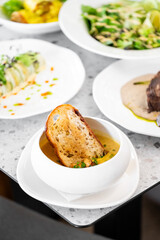 A rustic bowl of soup garnished with herbs, featuring a slice of toasted bread resting on the edge. The gourmet setting has an array of elegantly plated dishes in the background.