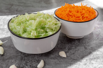 Two bowls filled with fresh chopped carrots and cucumbers stand on a gray background surrounded by garlic cloves, indicating preparation for a healthy dish