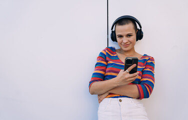 Confident buzzcut woman in striped shirt and white jeans standing casually with headphones and smartphone, smiling slightly while using mobile in front of clean white wall