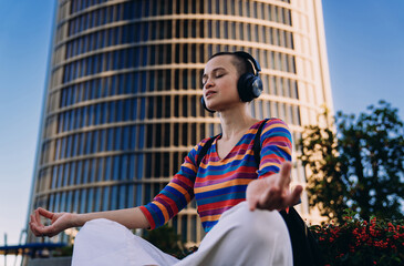 Young woman meditating calmly in lotus pose with headphones, sitting against modern skyscraper,...