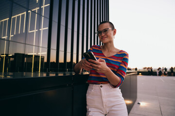 Caucasian woman in striped shirt and white jeans leans on rooftop terrace, smiling while using...