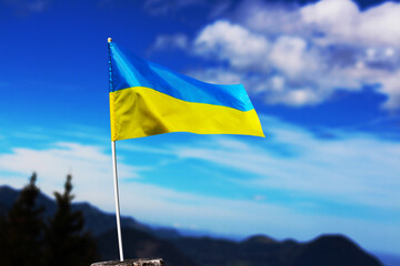 A Ukrainian flag waves against a bright blue sky with clouds and distant mountains in the background