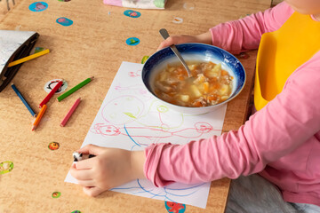 The child eats soup and draws with colored pencils at the children's table.