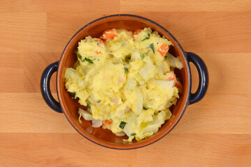 A top-down shot shows a small brown round baking dish with blue handles filled with mashed potato gratin on a wooden surface
