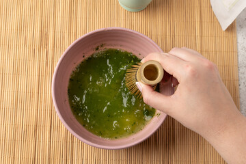 Woman whisking matcha green tea powder in bowl with bamboo whisk on bamboo mat