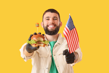 Young man in black gloves holding tasty burger and USA flag on yellow background