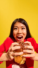 Closeup of joyful Asian woman expressing excitement holding big burger against bright yellow...