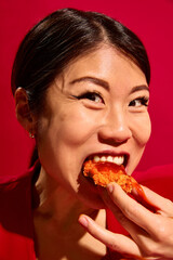 Close-up portrait of young excited Asian woman biting crispy chicken dripping with sauce against...