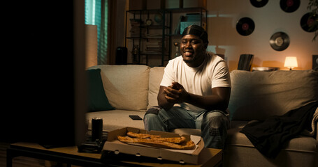 Relaxed man talks to a friend via video call in front of the TV. He laughs out loud, makes a heart gesture, and enjoys friendly gossip in a cozy evening moment.