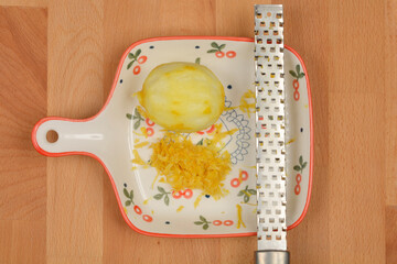 A small, decorative plate holds a peeled lemon and a pile of lemon zest, next to a metal zester, all arranged on a wooden cutting board