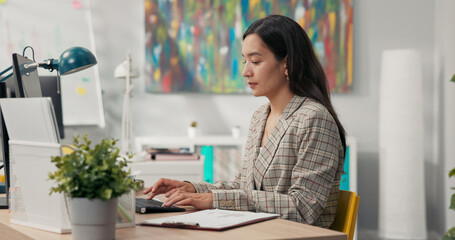 Attractive, experienced secretary of company is sitting in office in front of computer tapping...