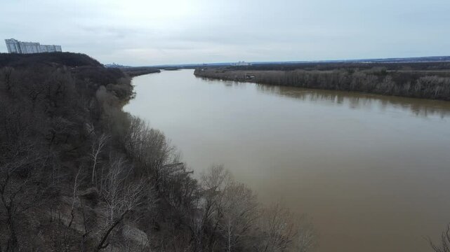Aerial footage showcases a serene river winding through vibrant forests under a cloudy sky, with distant modern buildings contrasting natures tranquility and urban encroachment